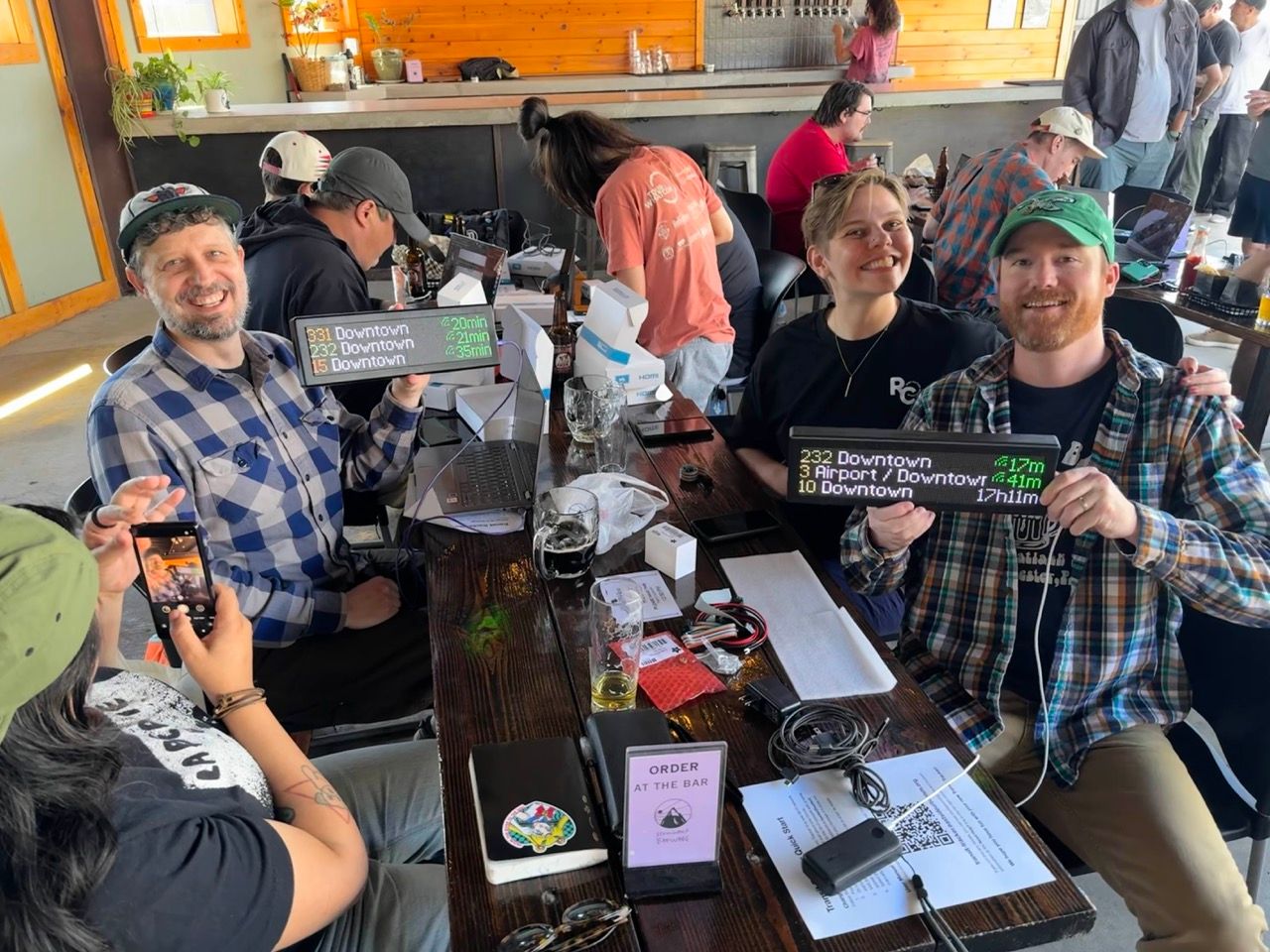 A group of people at a long table in an outdoor environment. Two people are smiling and holding up Transit Trackers.