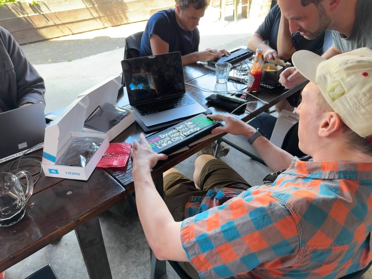 A person at a long table working on a Transit Tracker by seating the display. Several other people are in the background working on their Transit Trackers as well. One person is leaning over, apparently helping.