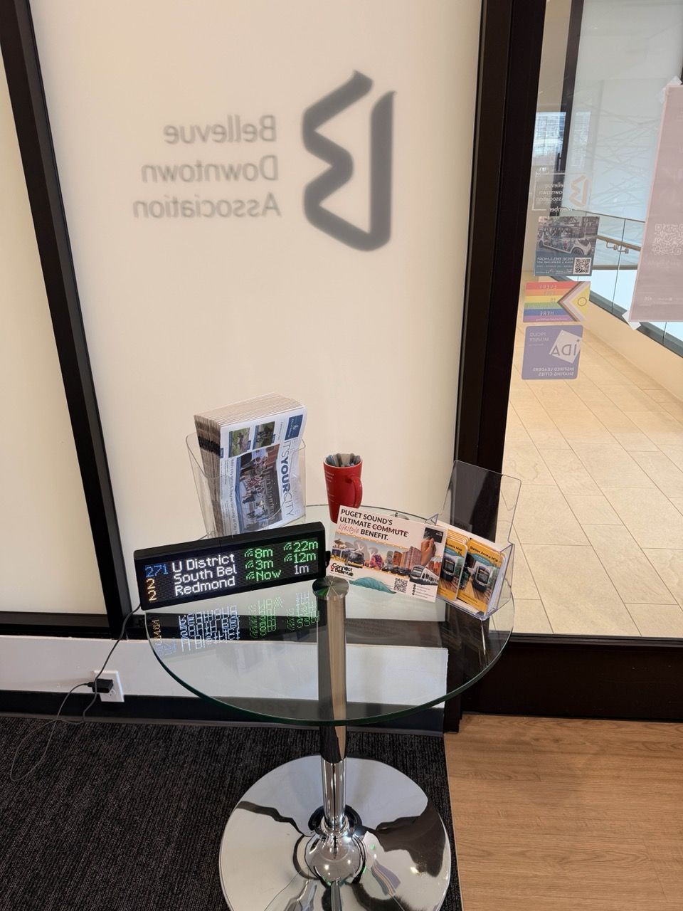A Transit Tracker on a glass table in front of a wall with "Bellevue Downtown Association" printed in reverse behind. The Transit Tracker is surrounded by various pamphlets and other information.