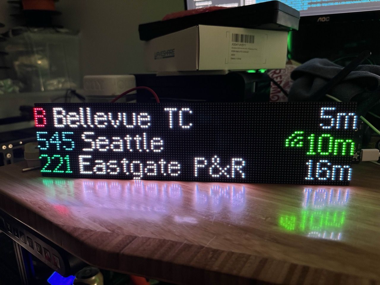 A bright LED matrix display on a desk displaying departure times for three different routes: B to Bellevue TC, 545 to Seattle, and 221 to Eastgate P&R. The desk is messy with various boxes and components strewn around.