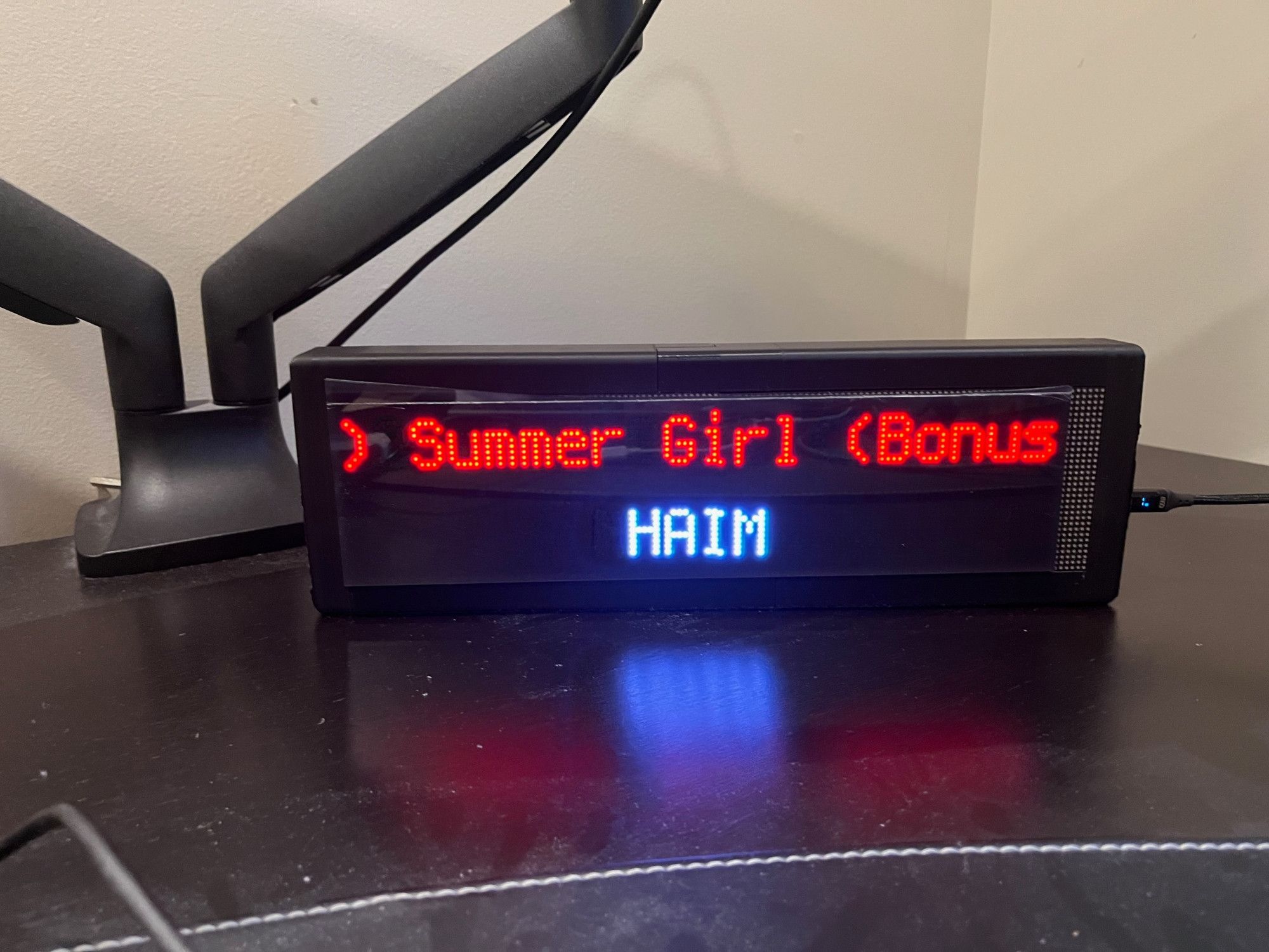 A picture of an LED matrix display in a 3D printed case sitting in front of a monitor arm on a desk. It is scrolling the song title Summer Girl in the top row and the band name, HAIM, in the bottom row.