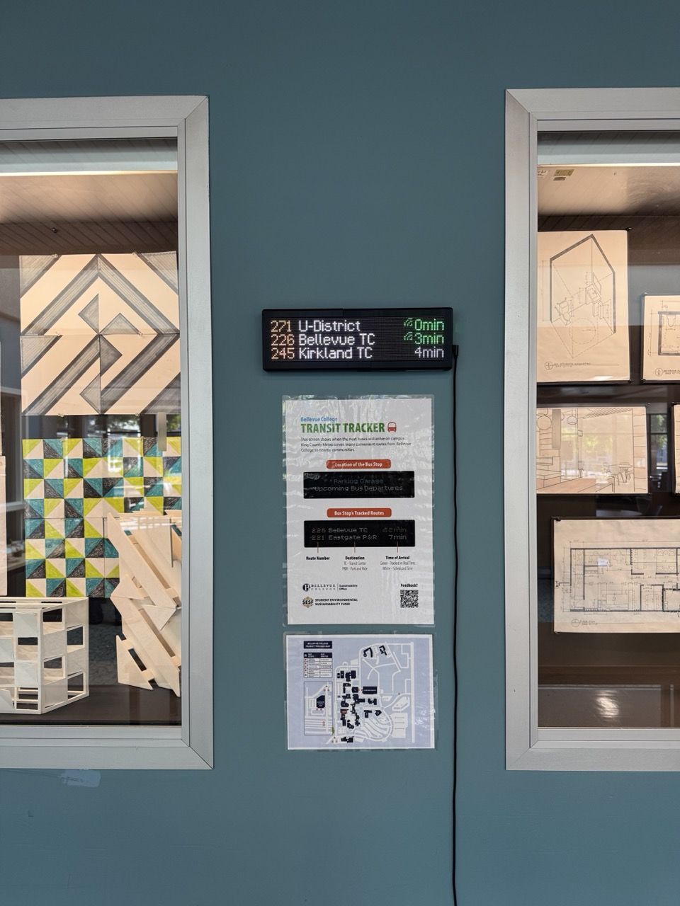 A Transit Tracker mounted on a wall with an instructional flyer and transportation map directly below. It is flanked by two windows.