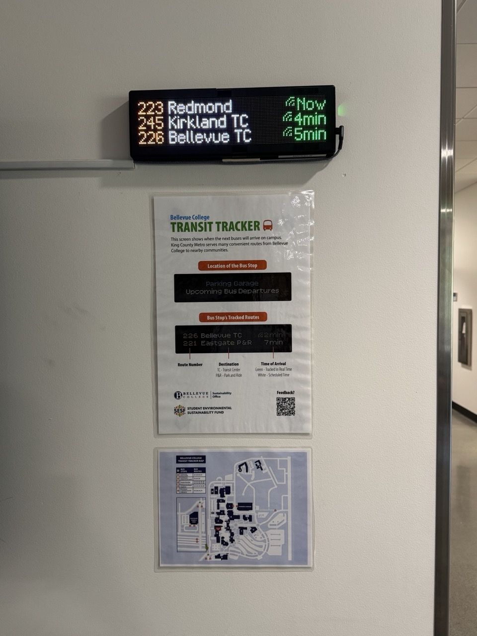 A Transit Tracker mounted on a wall in a hallway with an instructional flyer and transportation map directly below.