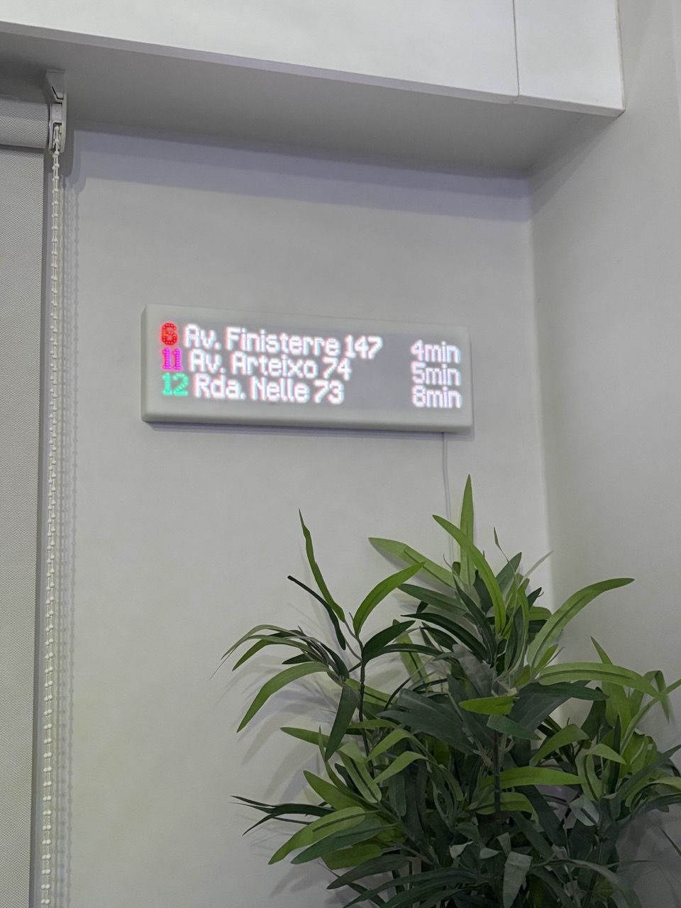 A Transit Tracker with a white frame and front plate mounted to the wall. The rest of the environment, including the wall and window shades, are very white as well. There is a plant below the Transit Tracker in the corner of the room.